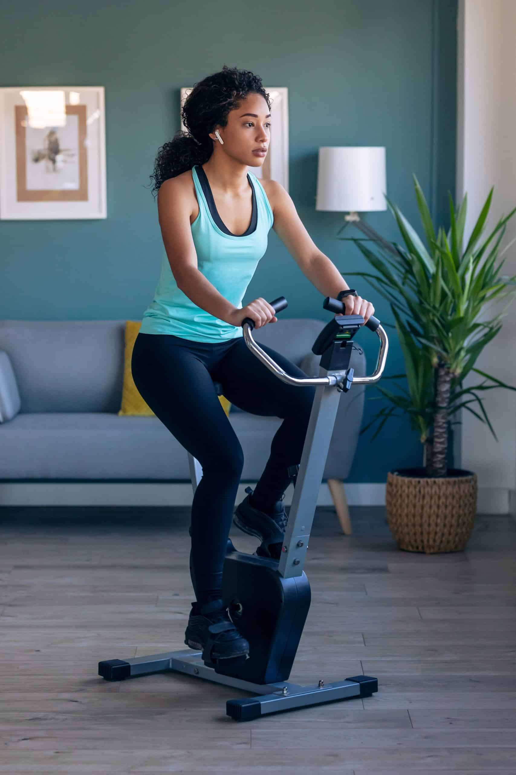 Sporty african young woman exercising on smart stationary bike and listening to music at home.